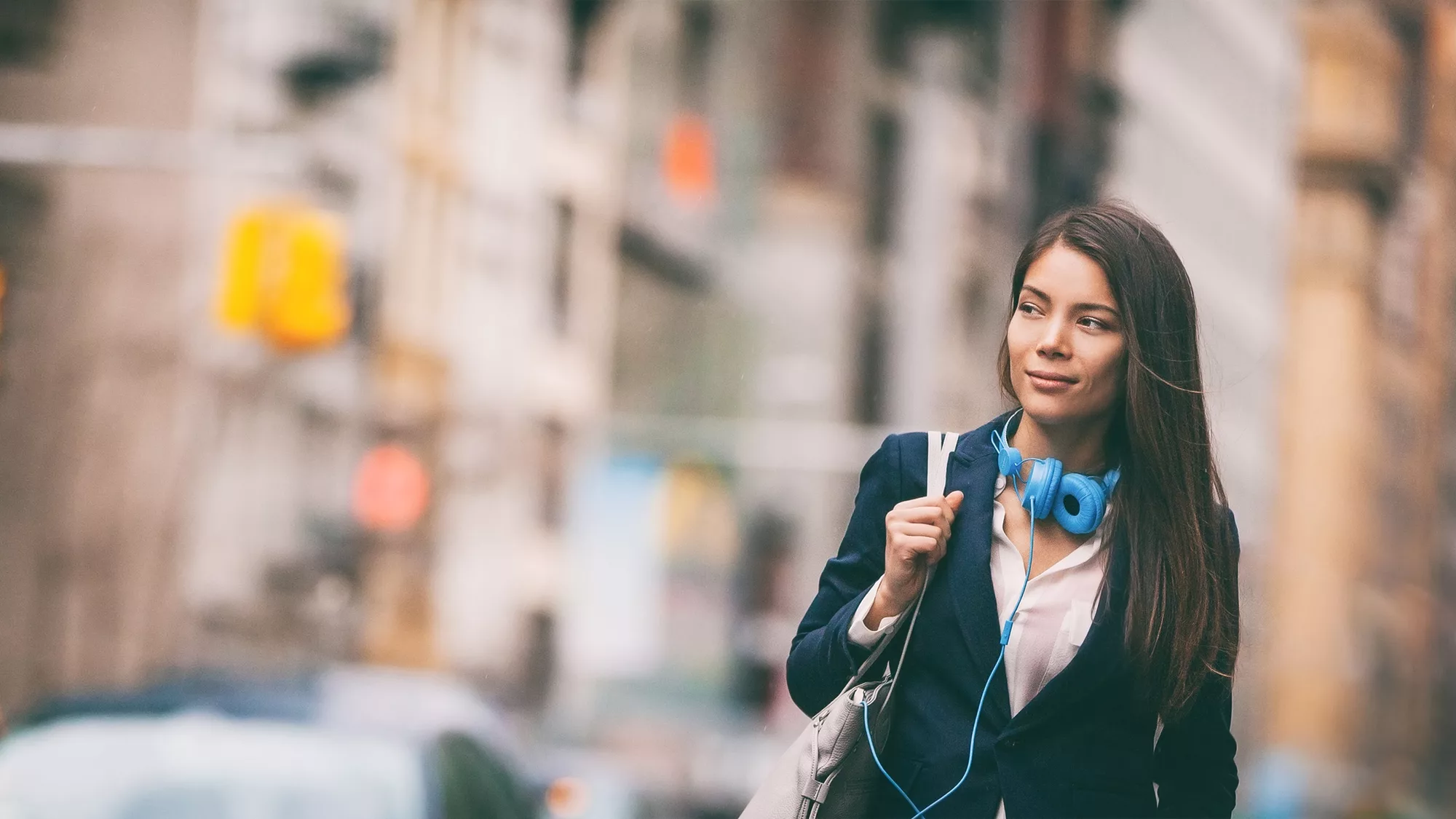 A young woman in a business suit walks in the city with blue headphones around her neck.